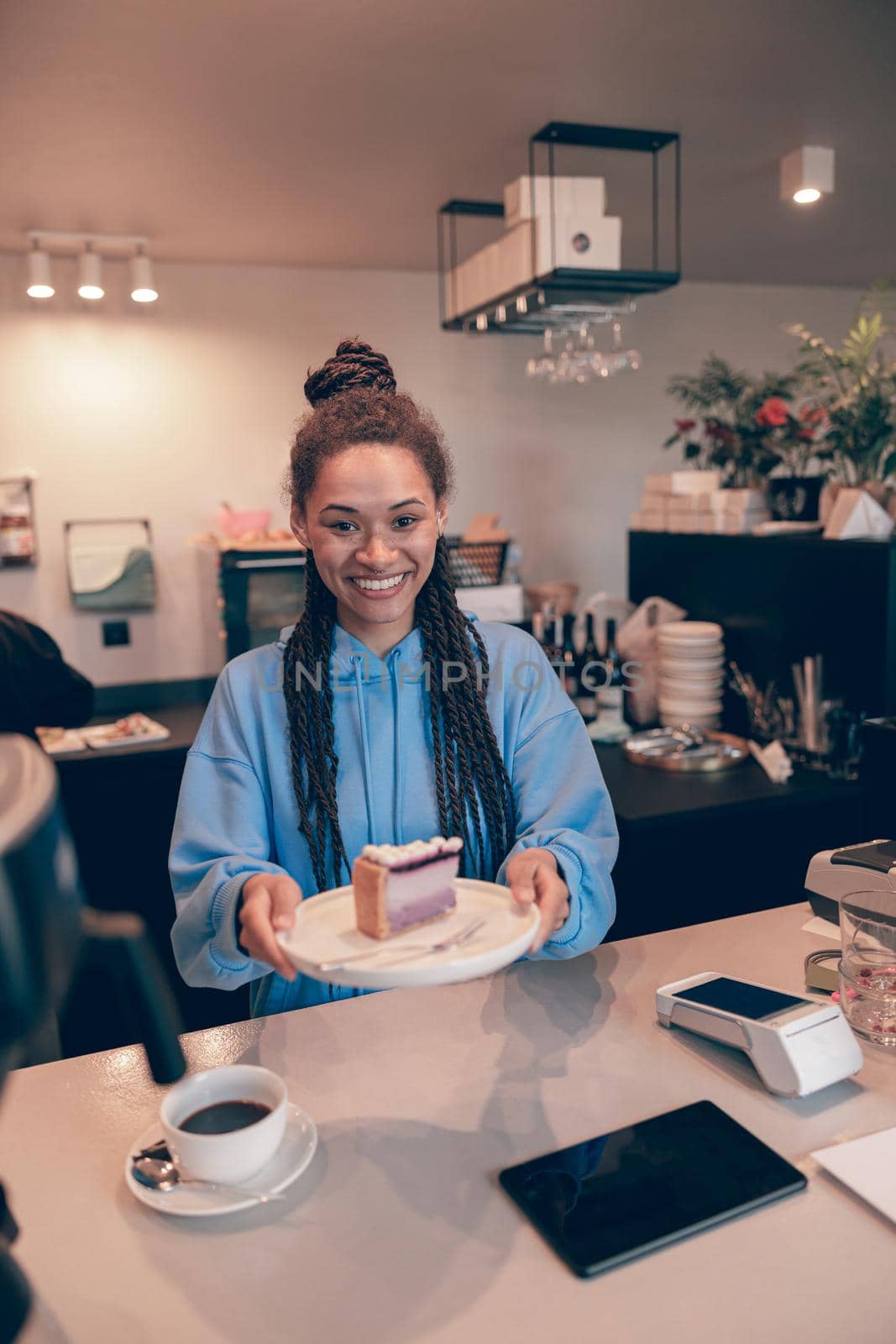 Portrait of young smiled female barista handing cheesecake on plate and coffee to camera. Vertical.