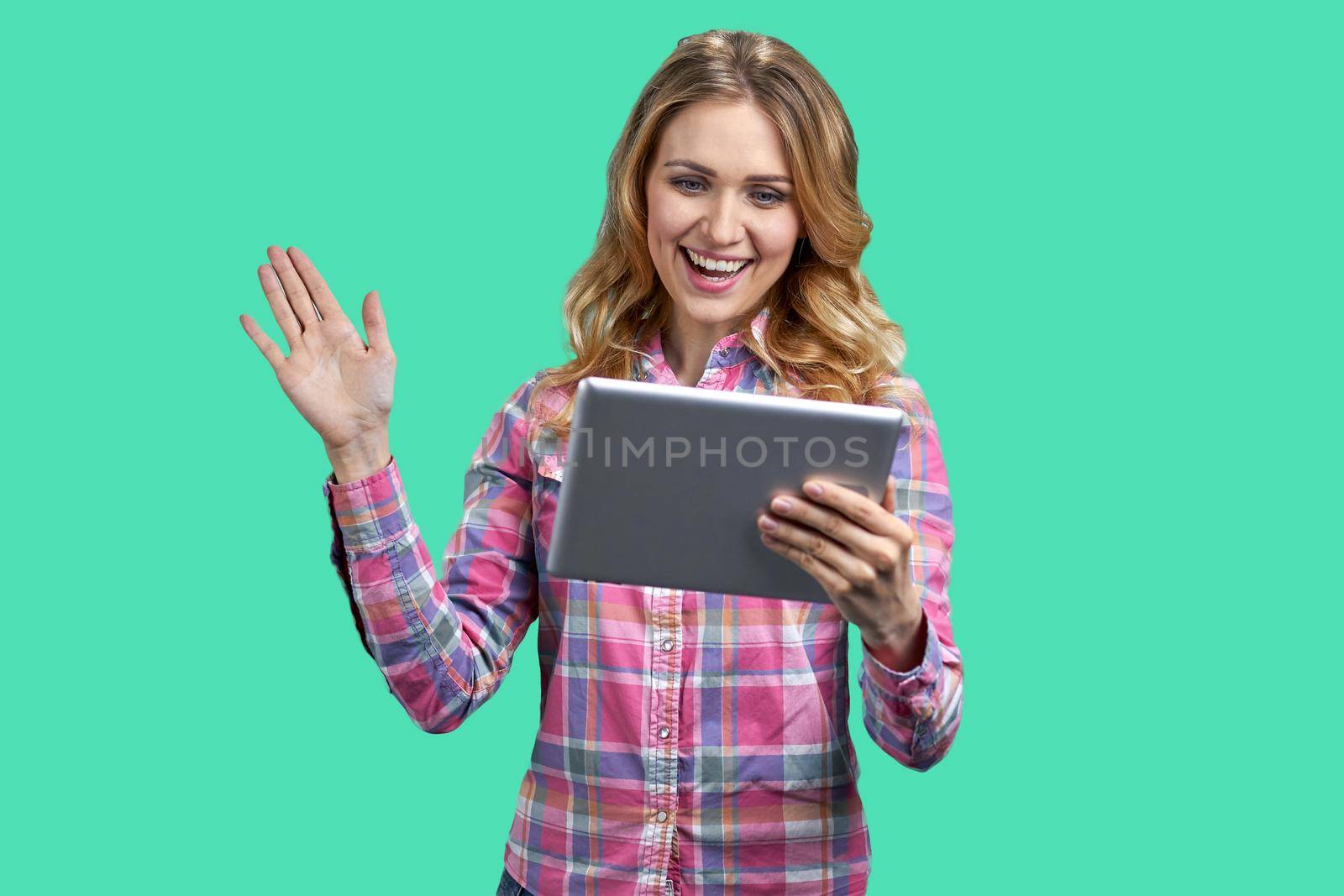 Young happy woman holding digital tablet and waving with hand. Young joyful woman having a video call on color background.