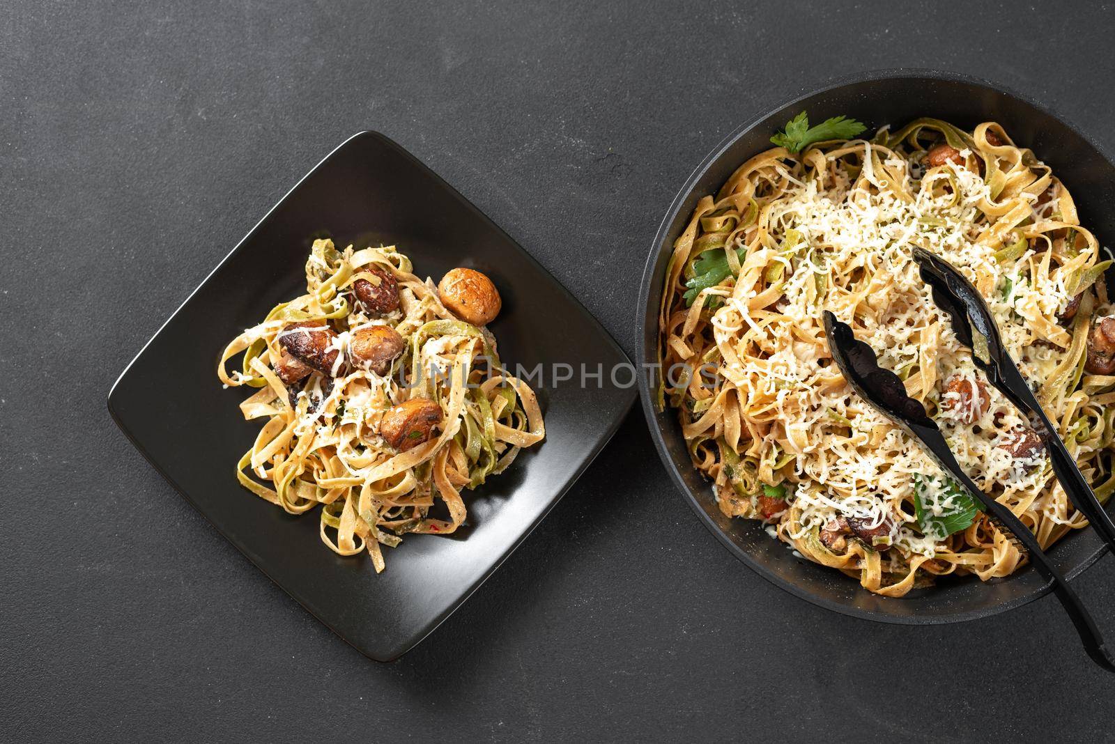 alfredo pasta with mushrooms in cream sauce on a black plate on a dark background. Freshly cooked pasta in a cast-iron skillet. Next to the plate with pasta is a classic Italian dish of fettuccini pasta