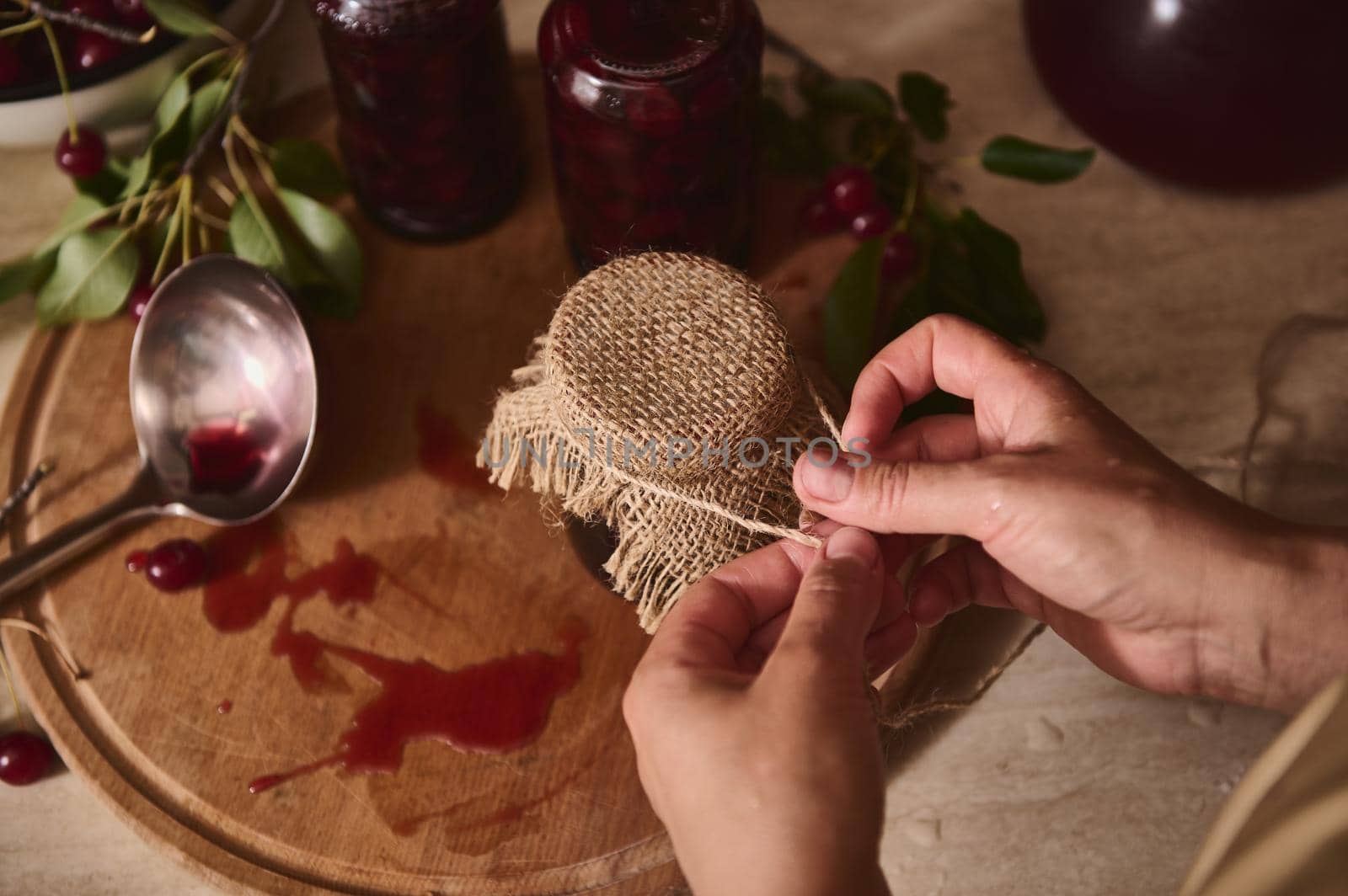 Top view of the hands of a pastry chef tying a rope on the burlap of the lid of a jar of freshly made cherry jam. A dirty ladle in the remnants of jam lies on a wooden board on the kitchen worktop