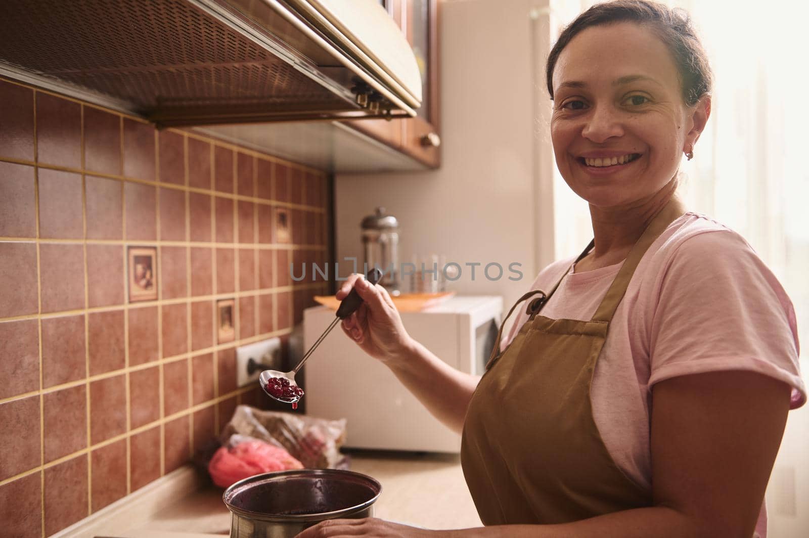 Gorgeous delightful multi-ethnic woman smiles toothy smile looking at camera while preparing a homemade cherry jam in the cozy rustic home kitchen. Food and drink concept, culinary