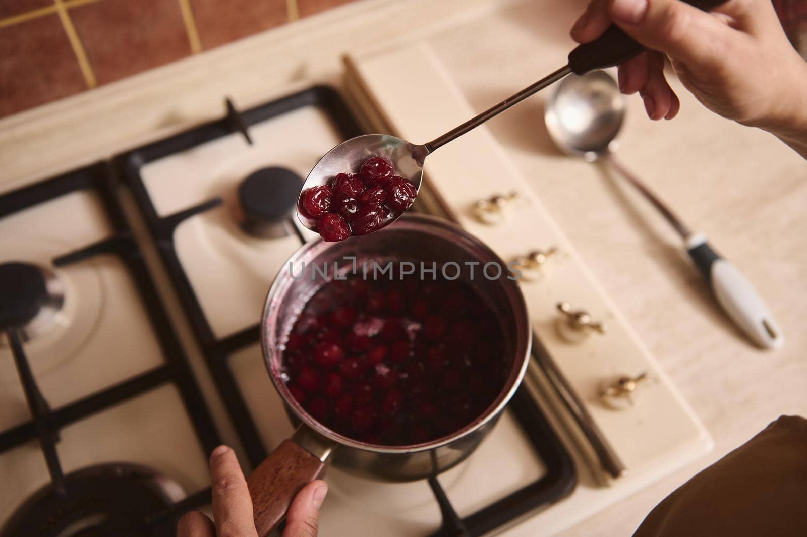 View from above of the hands of a chef confectioner standing by stove, using metal spoon stirring boiling jam of cherries with sugar while cooking homemade cherry berry confiture. Close-up