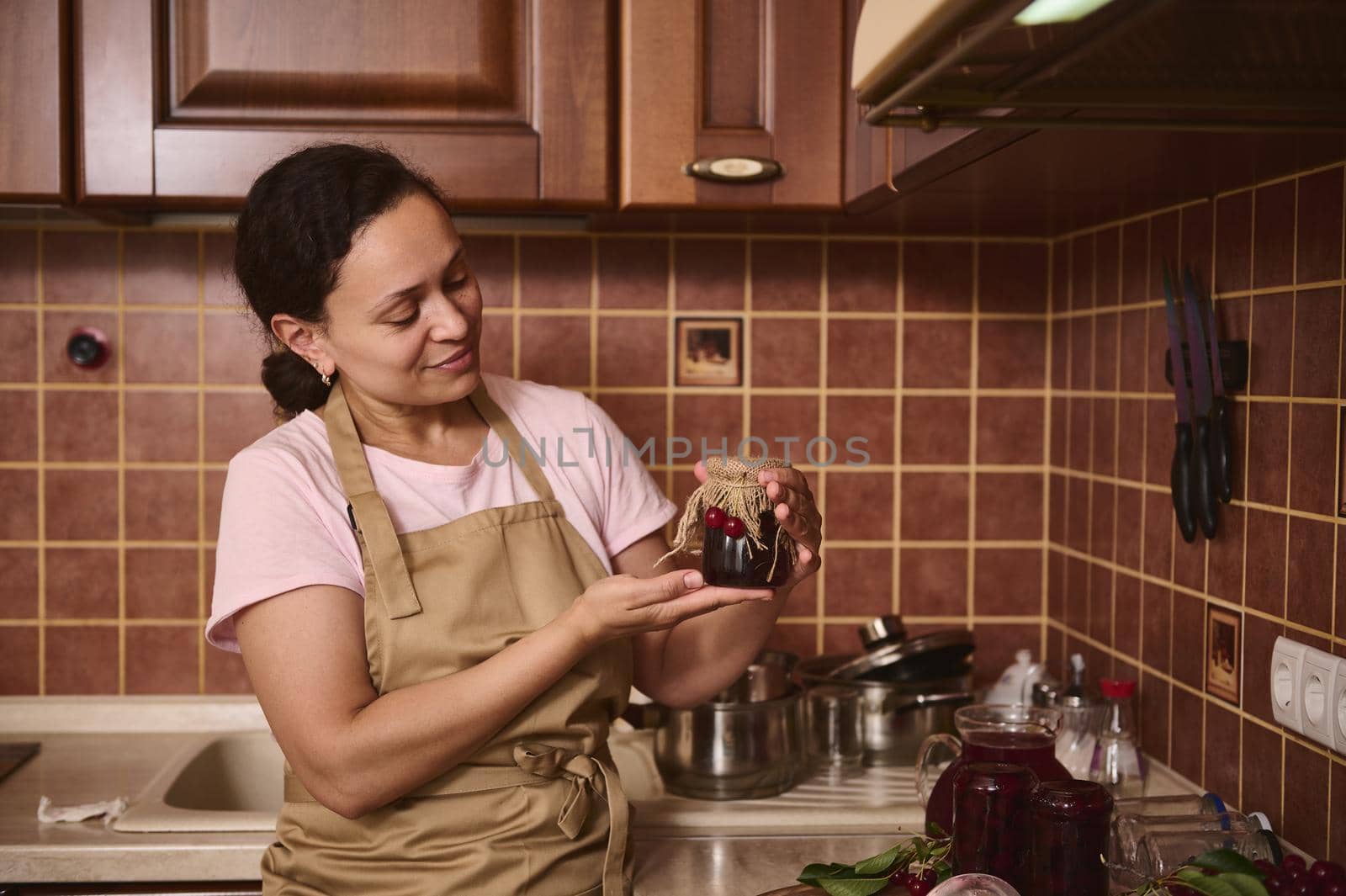 Charming African American woman, pleasant housewife wearing beige chef apron, holding a homemade cherry confiture jar with the burlap and cherries on the lid, standing at cozy rustic kitchen interior