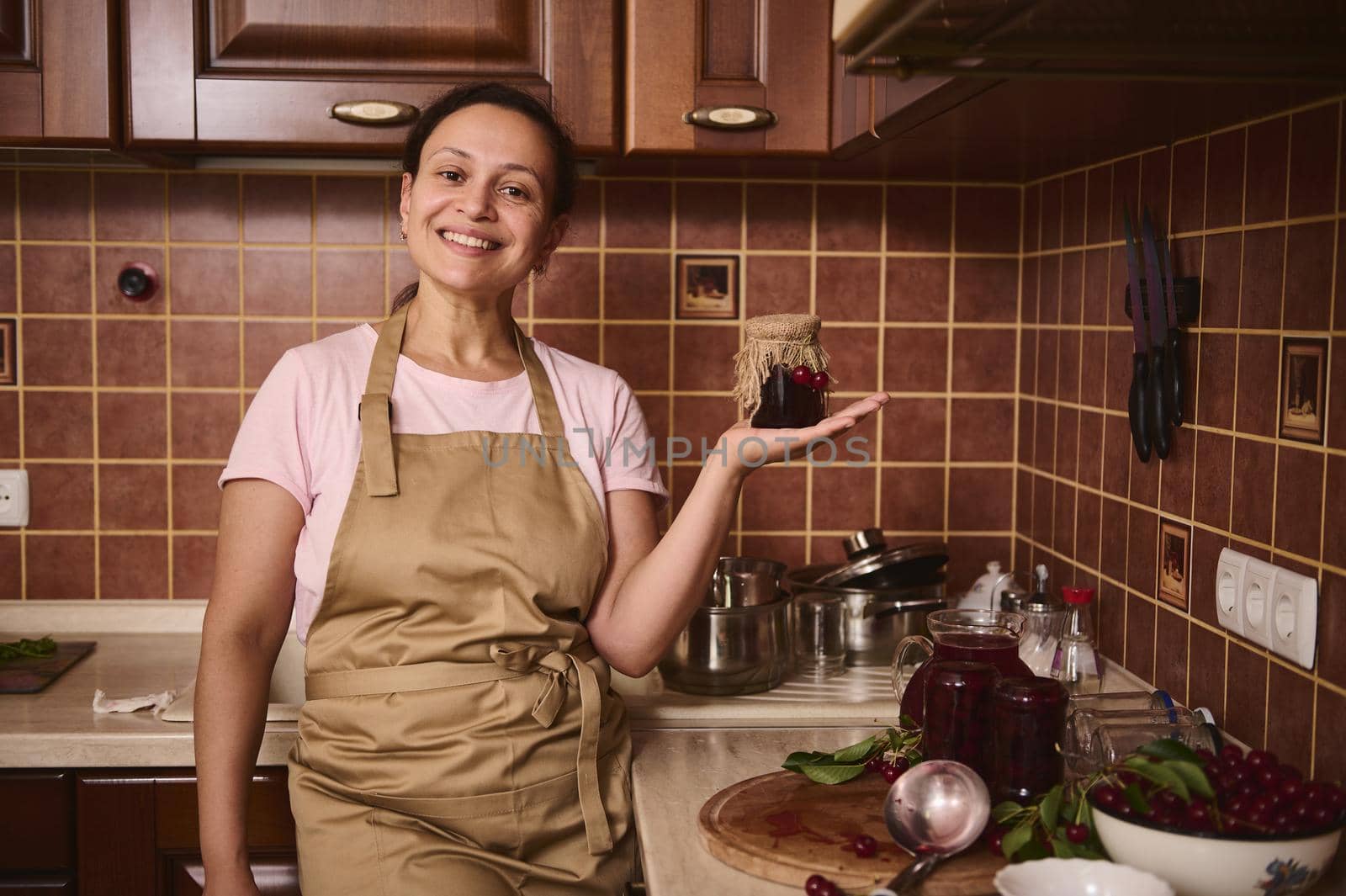Pleasant charming multiethnic woman confectioner, housewife in chef apron holds a jar of homemade cherry jam on her hand palm up and smiles, looking at camera, standing in a cozy rustic home kitchen