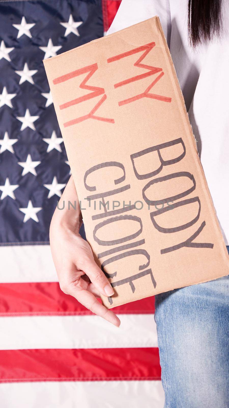 Young woman protester holds cardboard with My Body My Choice sign against USA flag on background. Girl protesting against anti-abortion laws. Feminist power. Equal opportunity Womens rights reedom