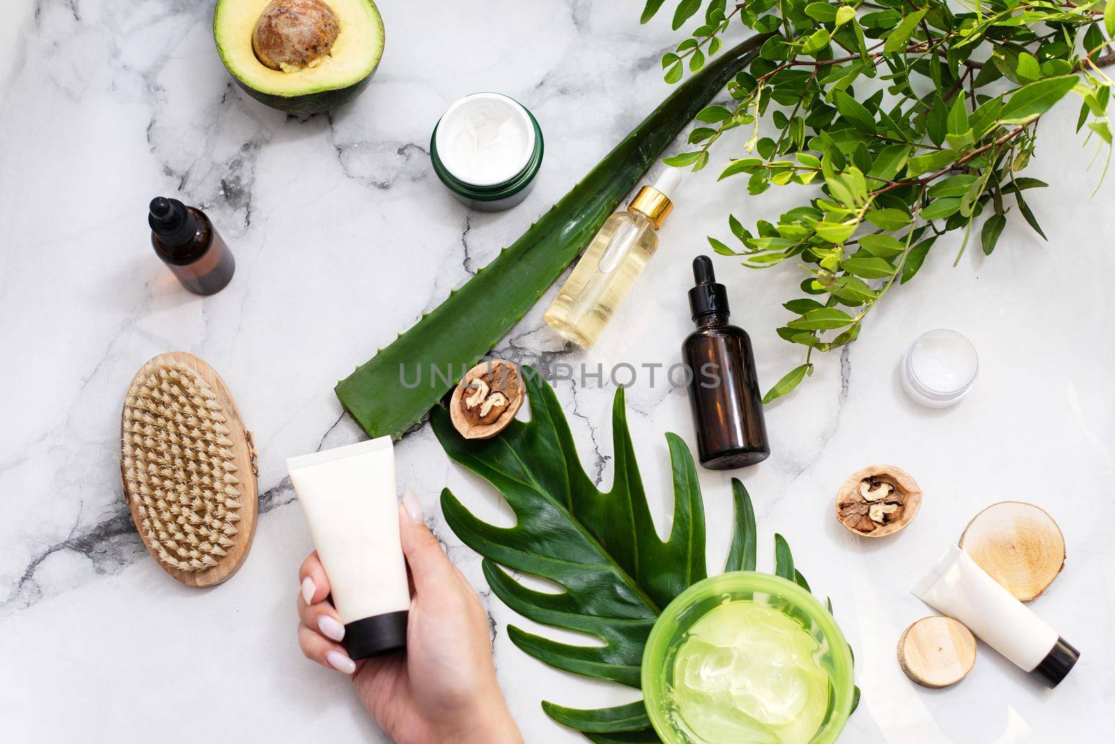 A woman's hand holds a moisturizer with a mockup on the background of a spa composition, tropical leaves, serum and gel