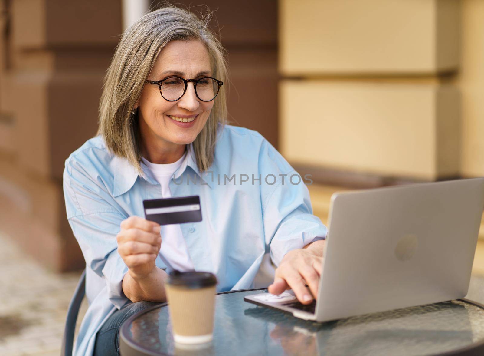 Charming mature european woman shopping or make online payment using laptop sitting outdoors street cafe terrace holding debit, credit card in hand. Mature woman happy vacation time traveling.