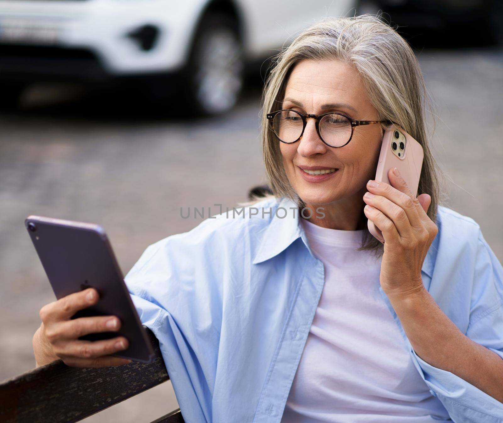 Enjoying free time mature grey hair woman talking on the phone reading news from digital tablet sitting on the bench at the streets of old european city. Mature woman answering call outdoors.