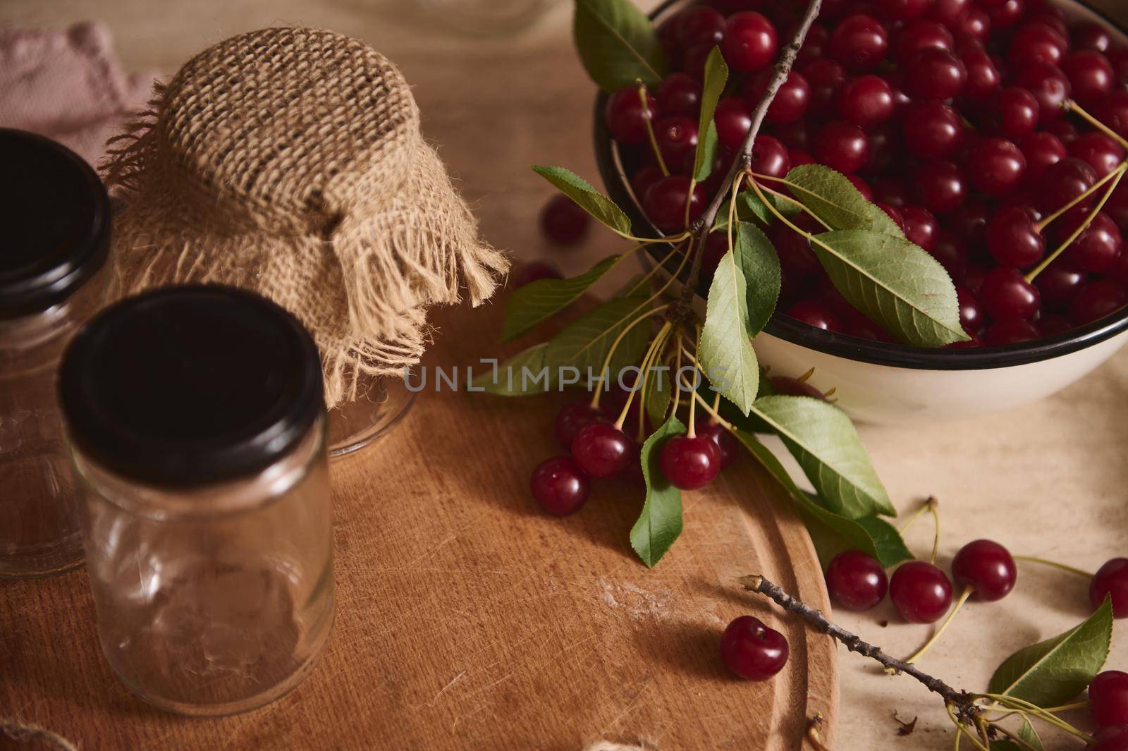 Close-up. Top view of sterilized glass jam jars with lids on a wooden board next to an enameled bowl of freshly picked cherries on a kitchen countertop. Preparation for conservation