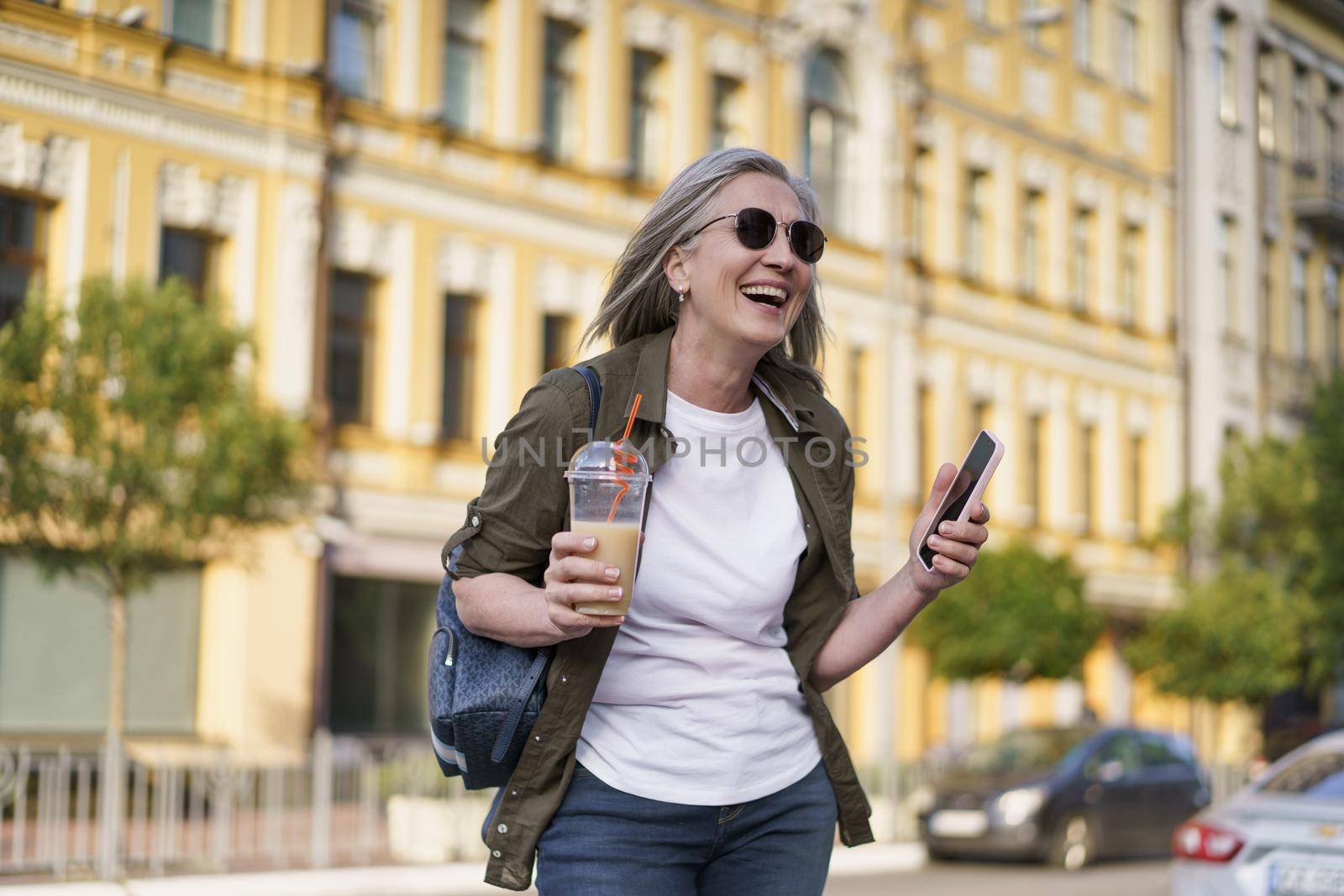 European senior woman with gray hair happy enjoying free time after work or traveling holding phone while having juice on the go using plastic cup in city background. Enjoying life mature woman by LipikStockMedia