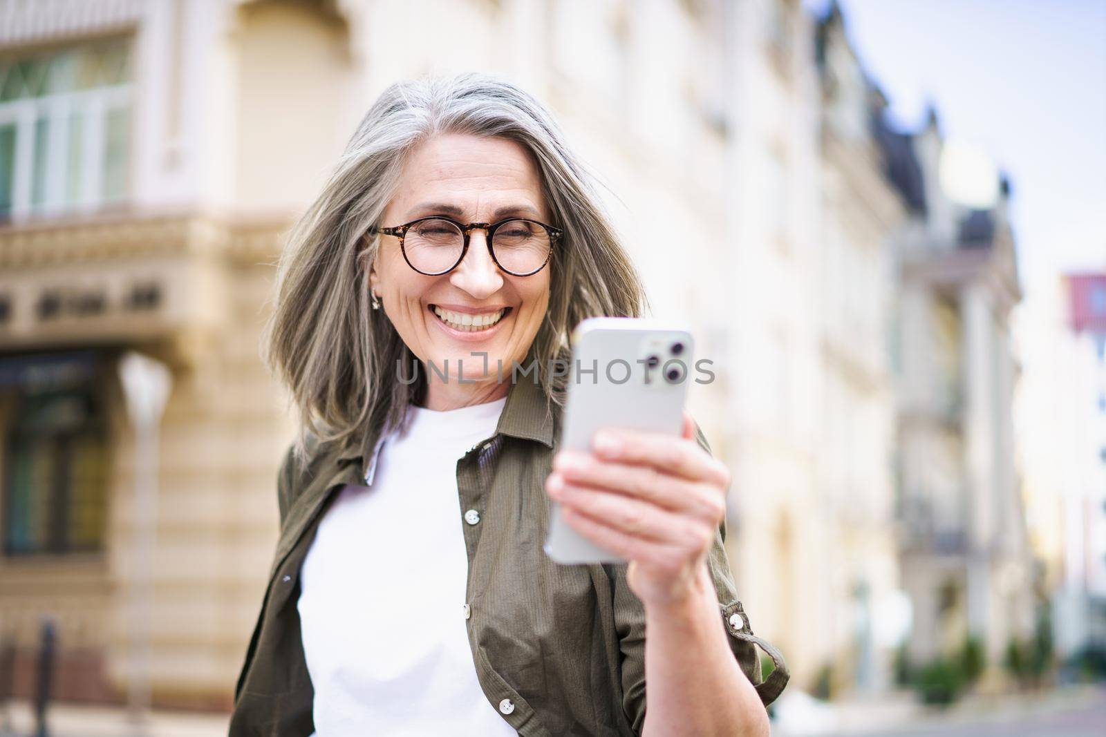 Beautiful mature woman with grey hair looking at white smartphone walking at the streets of old european town. Mature woman read text message or answering video call standing or walking outdoors.