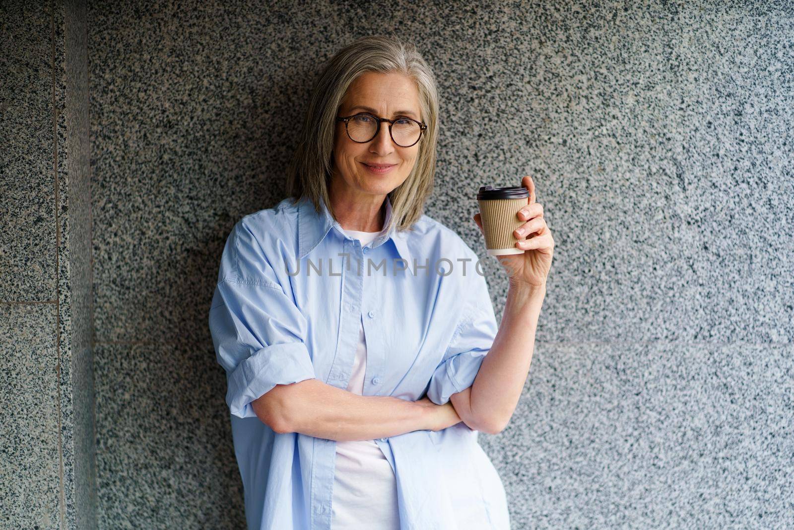 Charming mature grey hair woman at retirement time drink coffee using paper cup standing outdoors leaned on marble, granite wall smiling looking at camera with one arm folded.