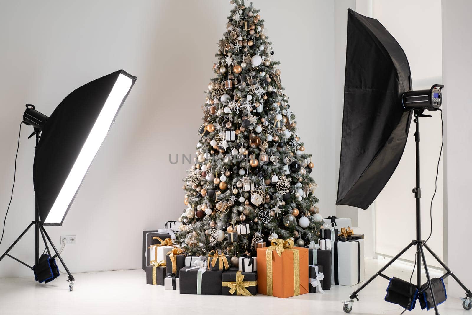 Christmas tree in white interior photo studio with stylish black and orange gift box standing like model with flash light by andreonegin