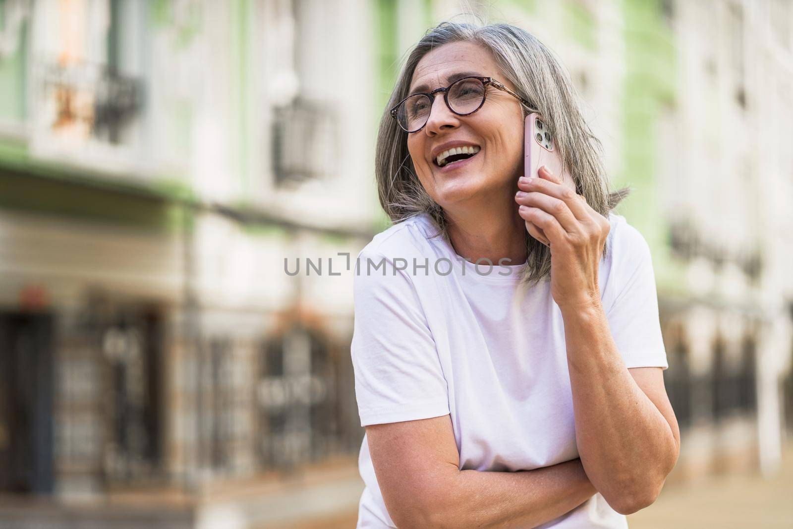 Charming mature business woman with grey hair talking on the phone holding smartphone standing outdoors of the streets of old urban city wearing white t-shirt. Silver hair woman outdoors by LipikStockMedia