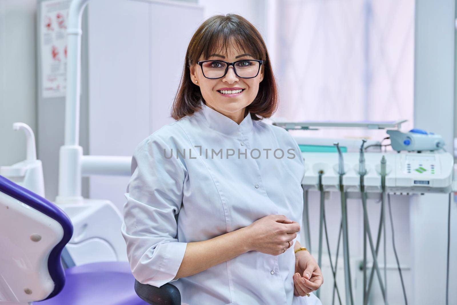 Portrait of smiling mature female dentist looking at camera in office. Positive middle aged doctor with glasses. Dentistry, medicine, health care, profession, treatment, stomatology concept