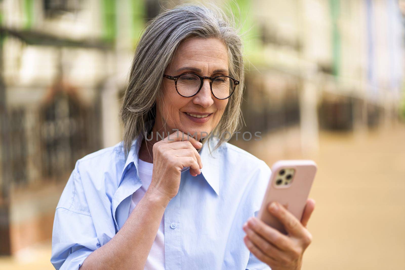 Charming mature woman with grey hair holding smartphone at the streets of old european town. Mature woman read text message or answering video call standing or walking outdoors by LipikStockMedia