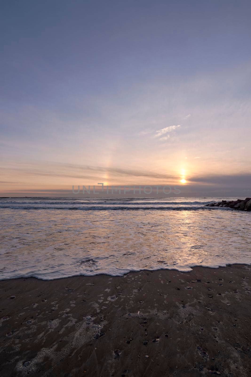 Sunrise from the beach with a breakwater. Rainbow, Circle around the sun, clouds and blue sky, waves, calm sea