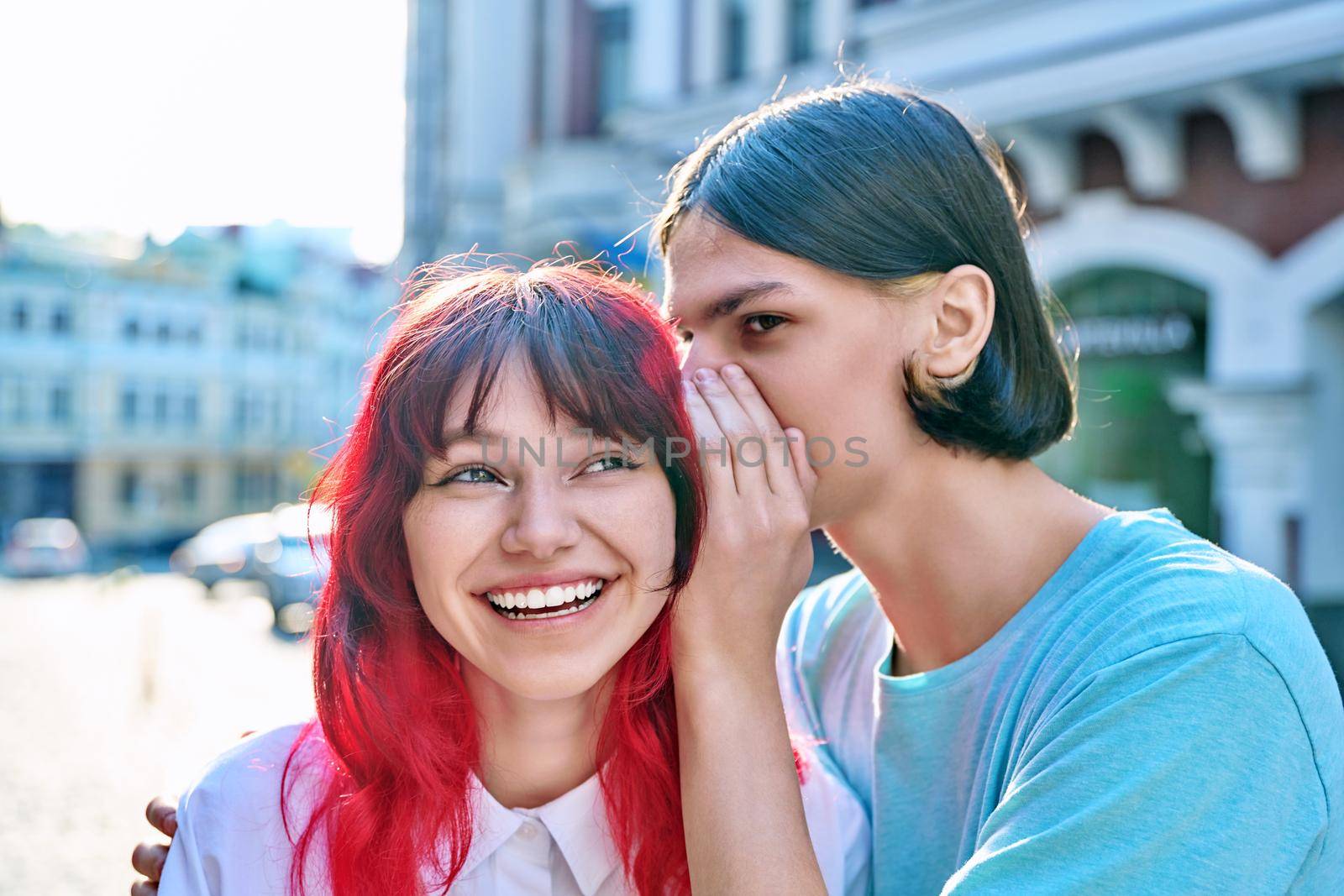 Couple of teenage students friends, guy whispers secret in ear of girlfriend on summer city street. Adolescence, friendship, relationships, surprise, emotions, youth concept