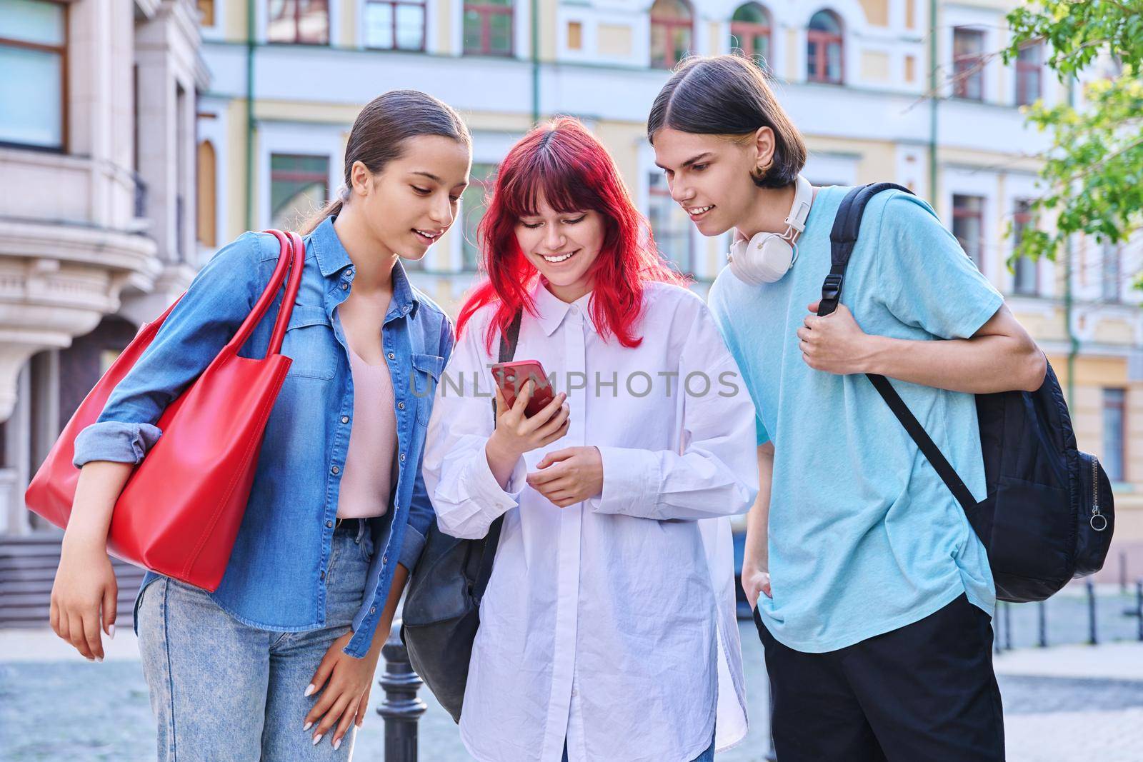 Three teenage friends met talking having fun outdoors. Smiling laughing teenagers with smartphone, together on city street. Communication, friendship, youth, lifestyle concept
