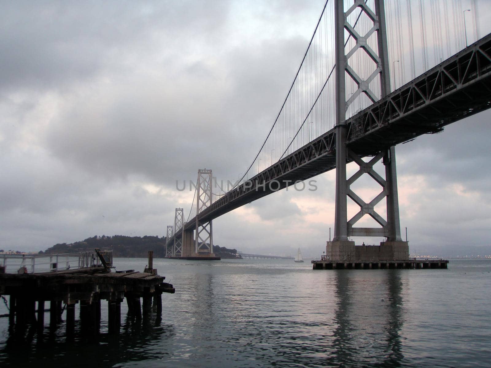 Old Pier and Bay Bridge on a Foggy Day with a sail boat in the background.