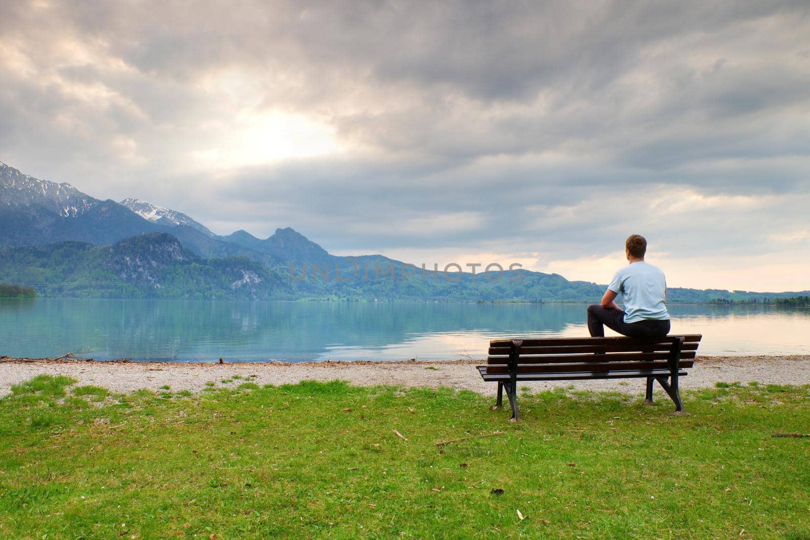 Tired adult man in blue shirt sit on old wooden bench at mountain lake coast by rdonar2