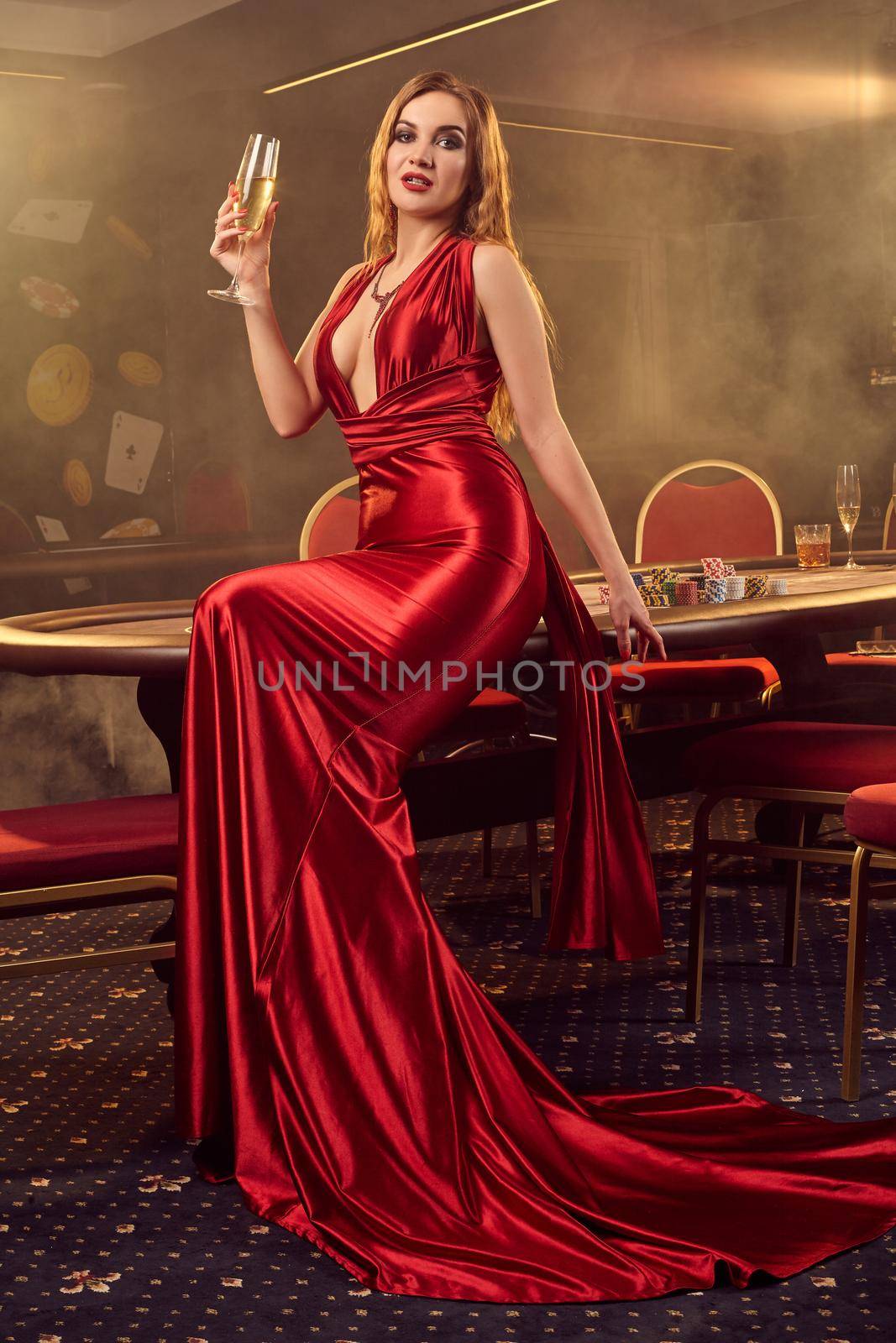 Young blond female in a long red satin dress, with a glass of champagne in her hand is posing sitting on a poker table and looking at the camera in luxury casino. Passion, cards, chips, alcohol, win, gambling - it is a female entertainment. Smoke background.