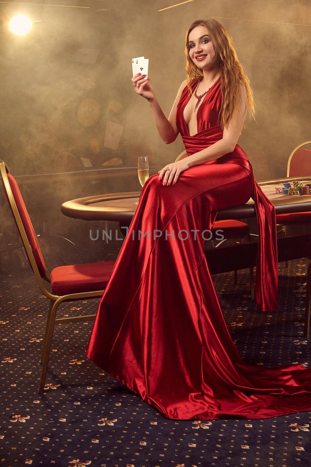 Young blond maiden in a long red satin dress, with two aces in her hand is smiling widely and posing sitting on a poker table in luxury casino. Passion, cards, chips, alcohol, win, gambling - it is a female entertainment. Smoke background.