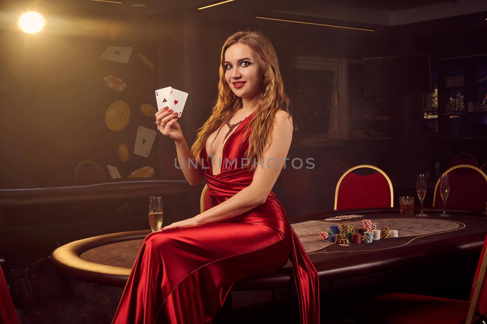 Showy blond lady in a long red satin dress, with two aces in her hand is posing sitting sideways on a poker table in luxury casino. Passion, cards, chips, alcohol, win, gambling - it is a female entertainment. Smoke background.