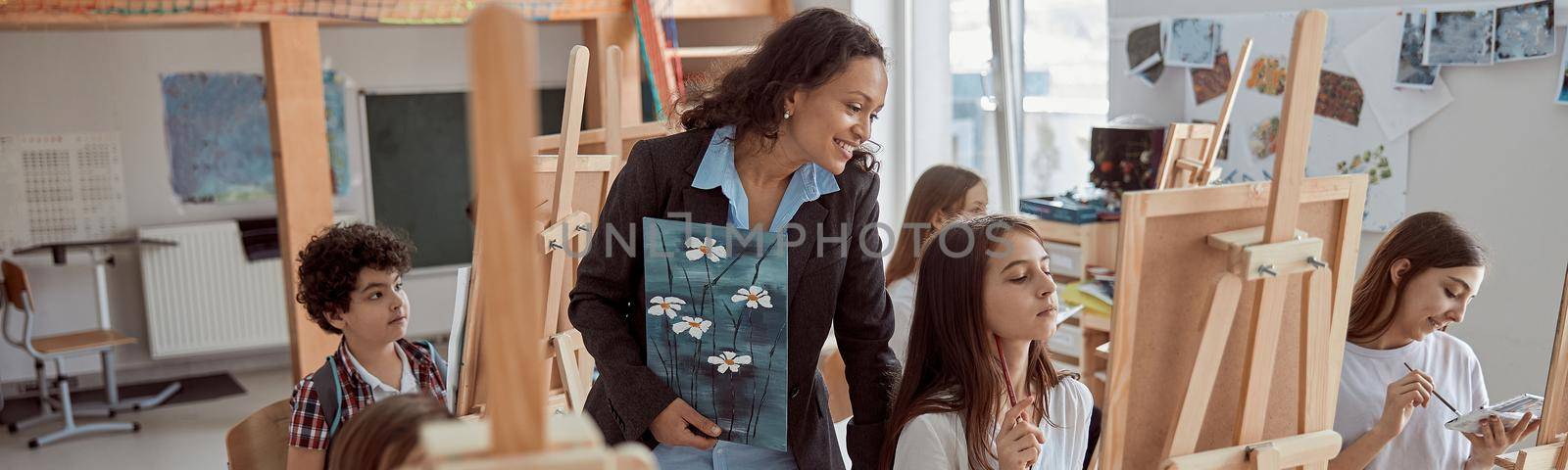 Young beautiful confident teacher is helping a kid to draw on a group lesson in a white modern minimalistic classroom.