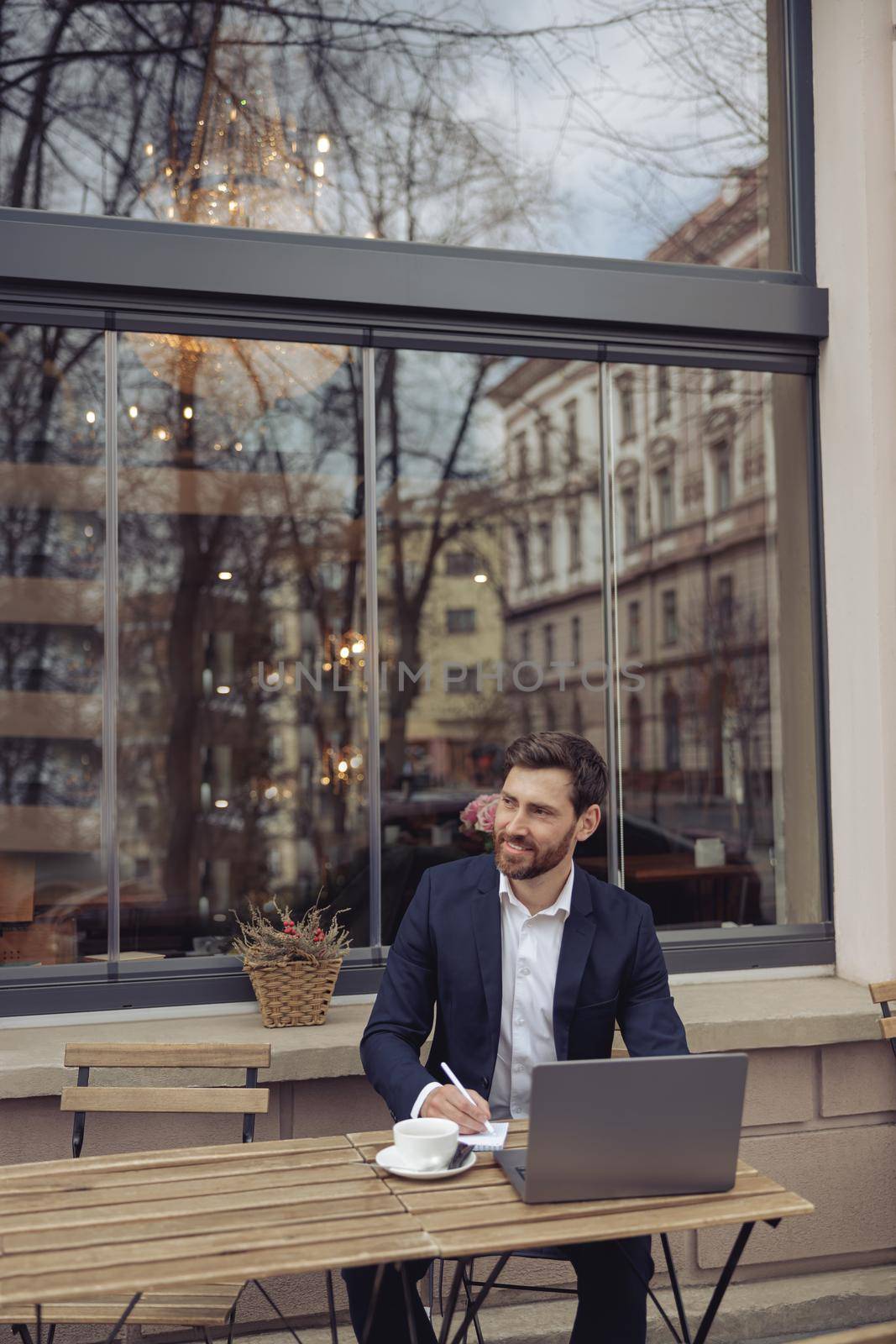 Attractive elegant man taking notes while working at laptop in cafe outside. Writer at coffee break.