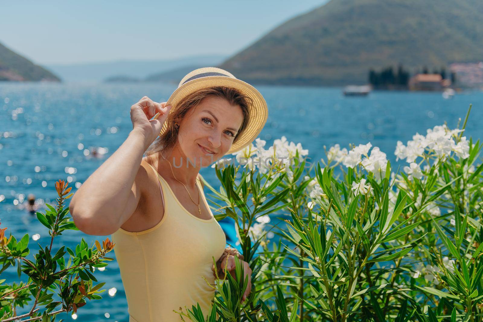 Summer photo shoot on the streets of Perast, Kotor, Montenegro. Beautiful girl in yellow dress and hat. smiling tourist girl with hat. Spectacular view of Montenegro with copy space. fashion outdoor photo of beautiful sensual woman with blond hair in elegant dress and straw hat and bag, posing in Montenego's city Perast by Andrii_Ko