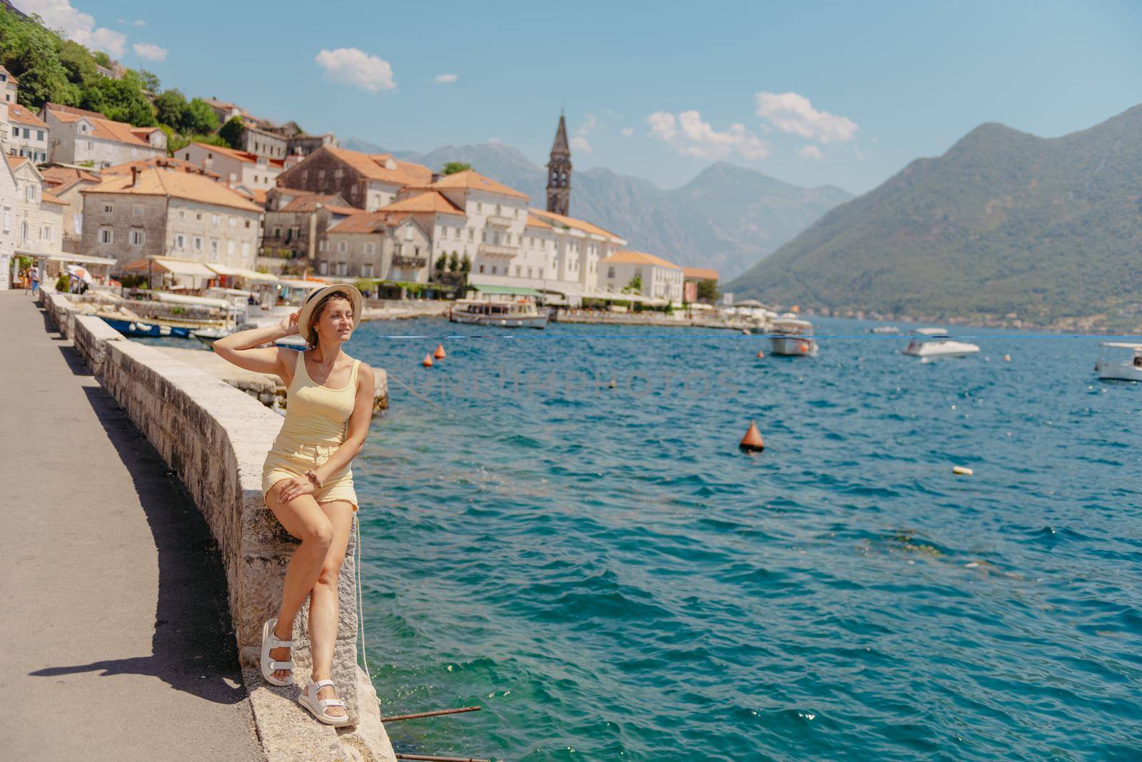 Summer photo shoot on the streets of Kotor, Montenegro. Beautiful girl in yellow dress and hat. smiling tourist girl with hat. Spectacular view of Montenegro with copy space. Ю fashion outdoor photo of beautiful sensual woman with blond hair in elegant dress and straw hat and bag, posing in Montenego's city Perast.