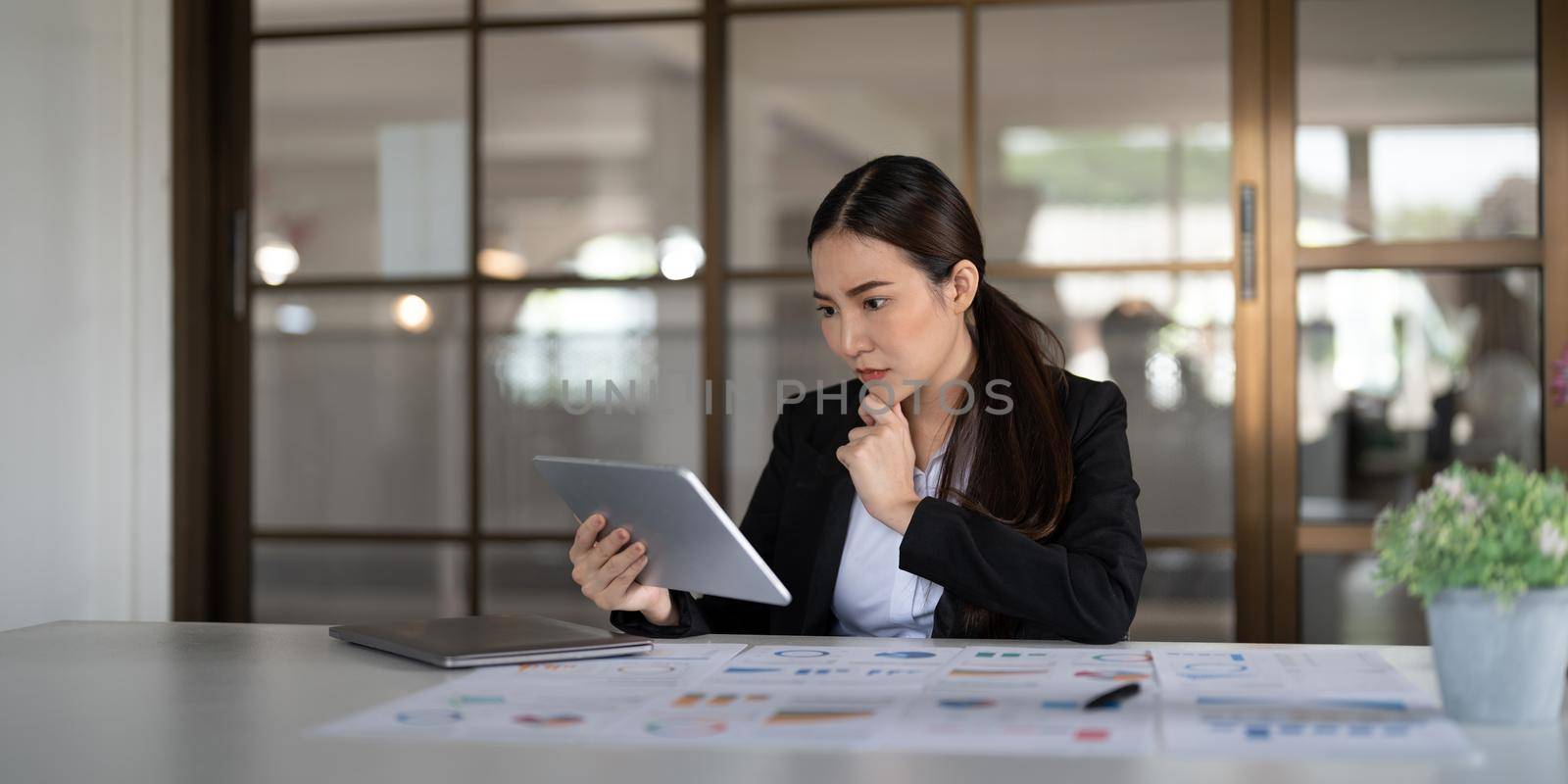 Portrait seriously business asian woman using digital tablet for do math analysis finance on wooden desk in office, tax, accounting, financial concept.