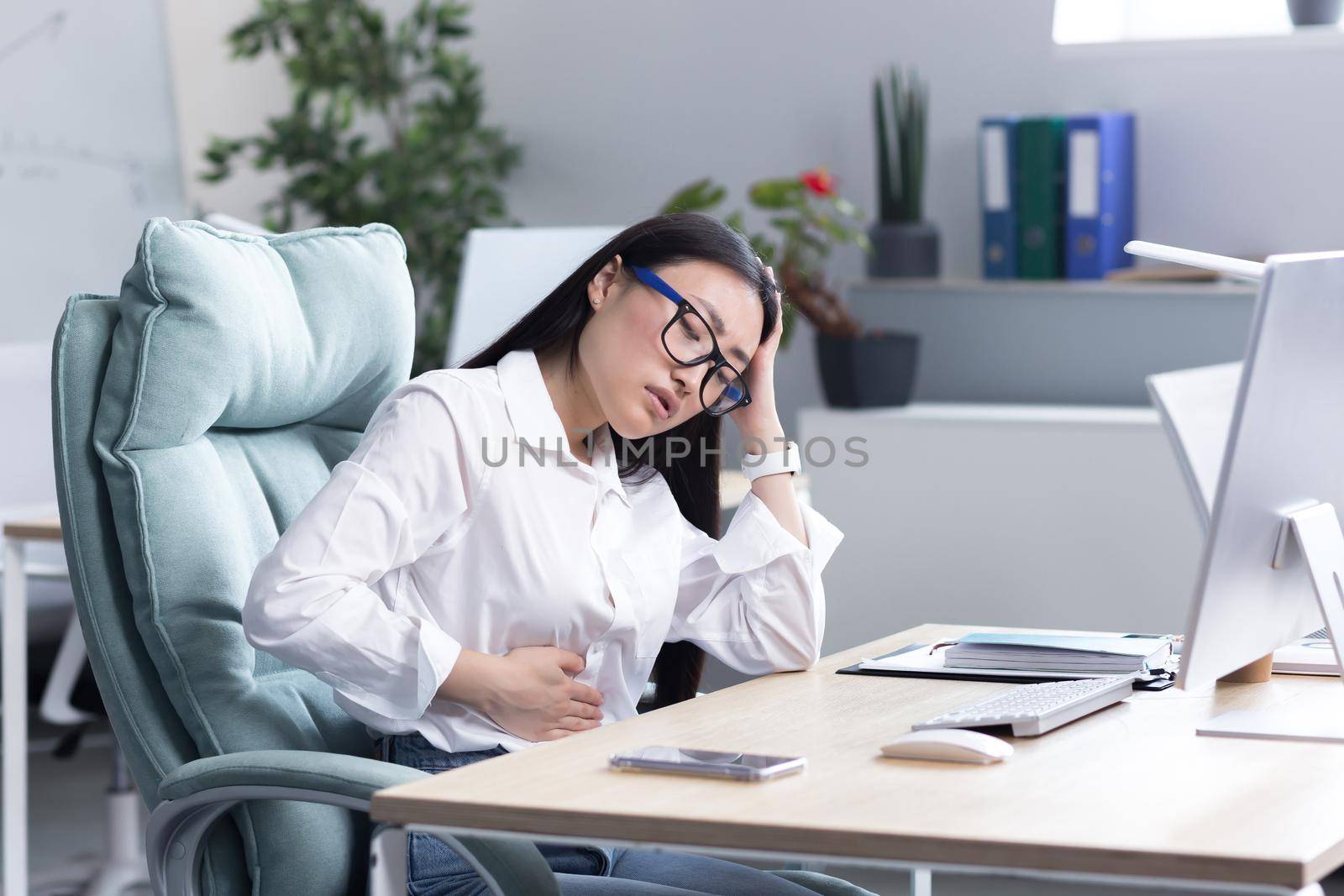 Abdominal pain at the workplace. A young beautiful Asian woman in glasses works in the office at the computer, feels pain in her stomach, holds her hand to her stomach, grimaced in pain.