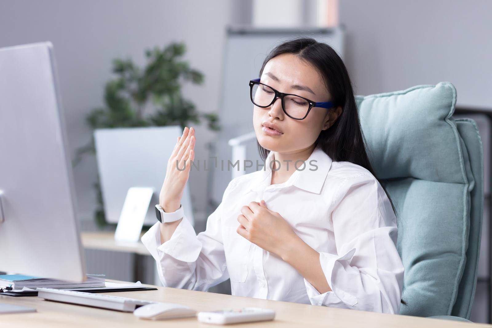 Close-up photo. Heat in the workplace. Young Asian woman sitting in the office at the desk waving her hand at her face, it's very hot, she feels bad, she needs fresh air, take a break.