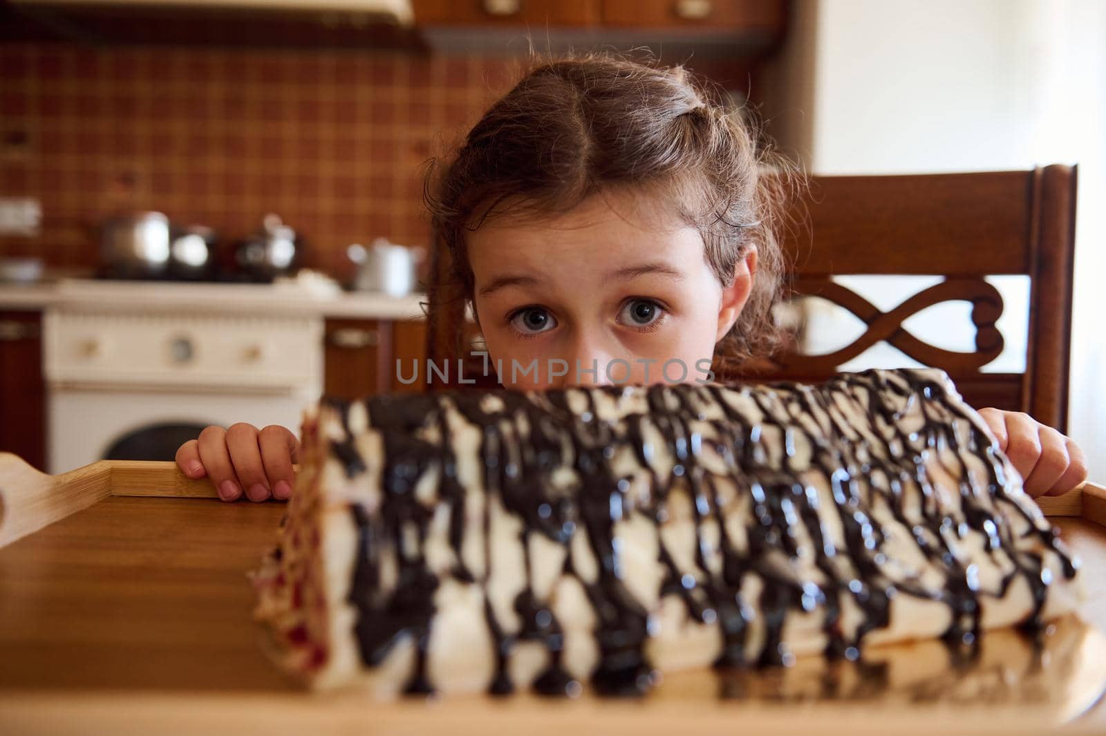 Portrait of adorable European little girl looking at camera from under blurred cherry chocolate pie - Monastery log at rustic cozy home wooden kitchen interior