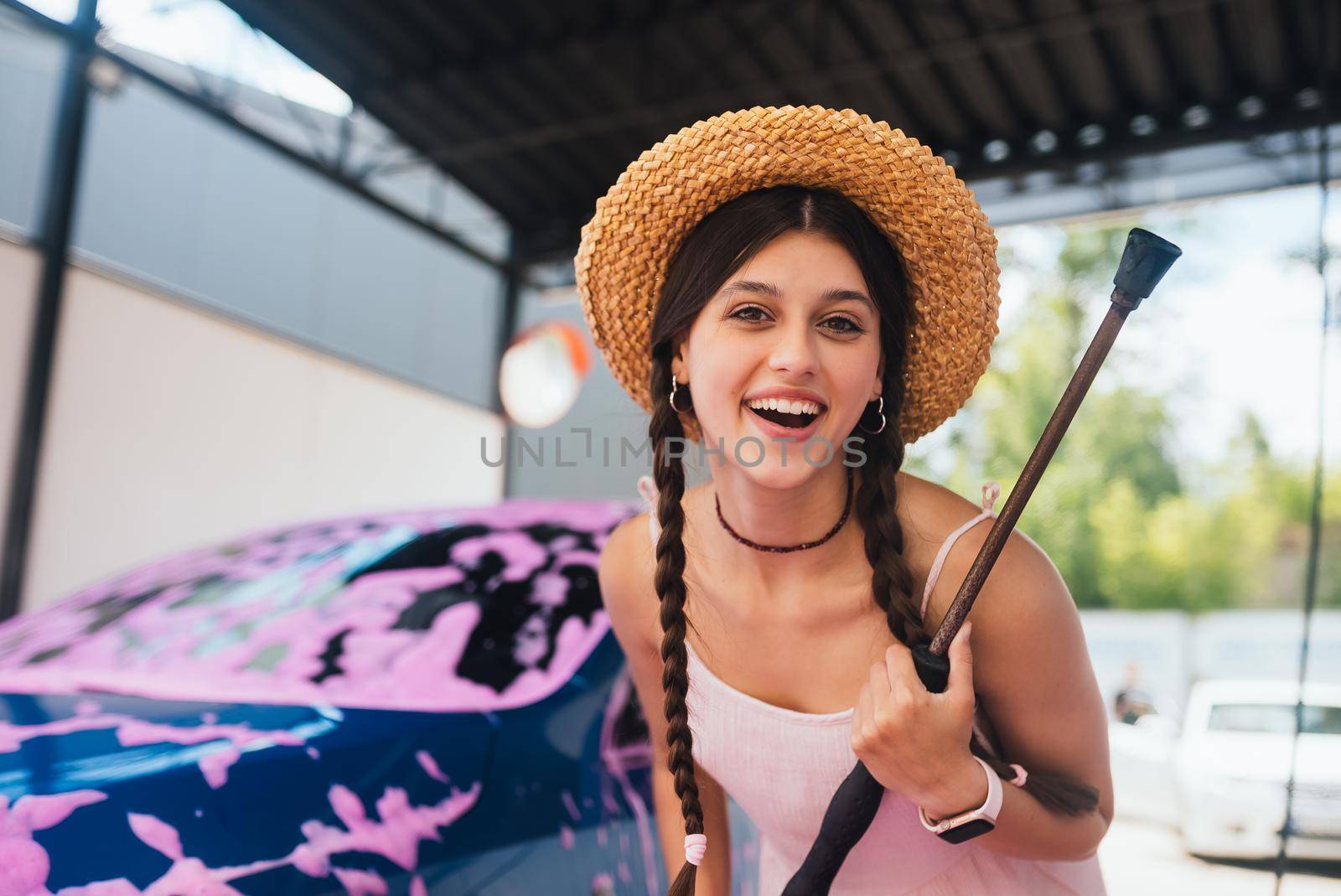 Young woman with high pressure hose stands by car covered in pink foam