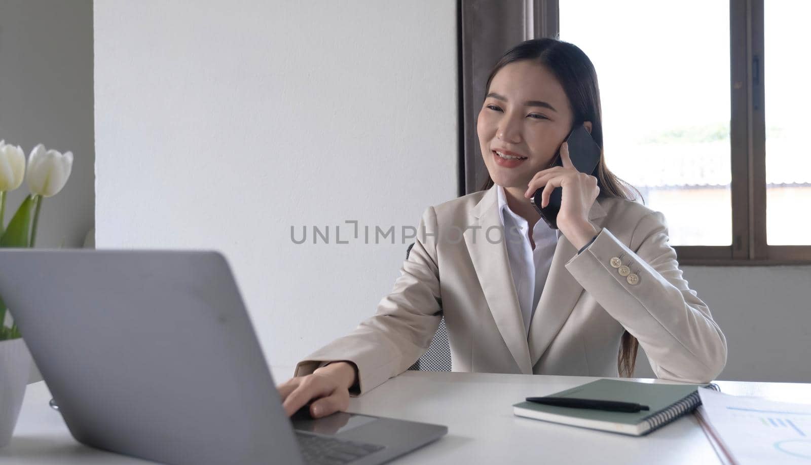 Young Asian woman talking on the phone using a laptop computer in the office. by wichayada