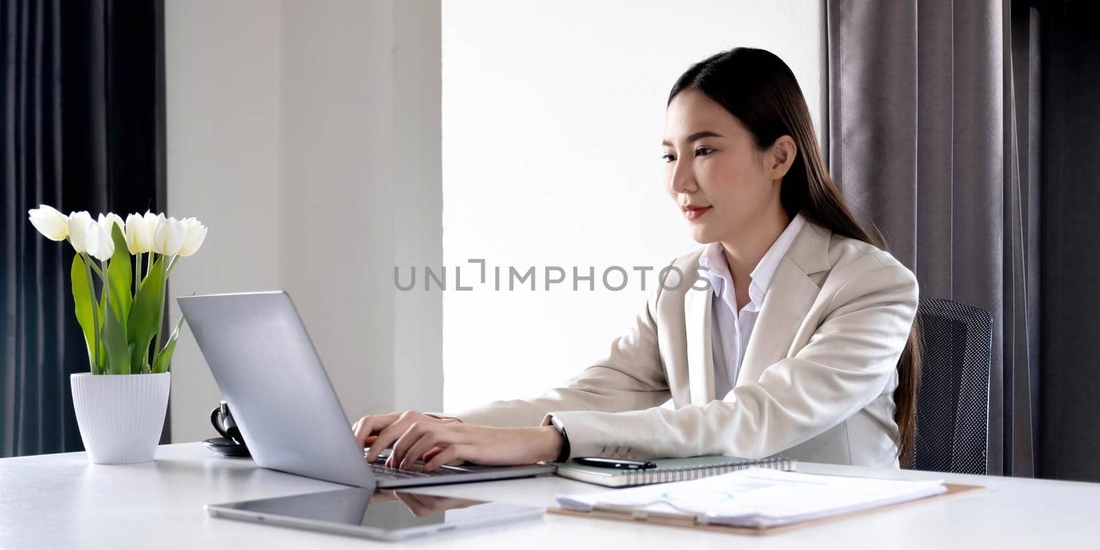 Front view of a beautiful Asian businesswoman working on a tablet coffee cup placed on the office table. by wichayada