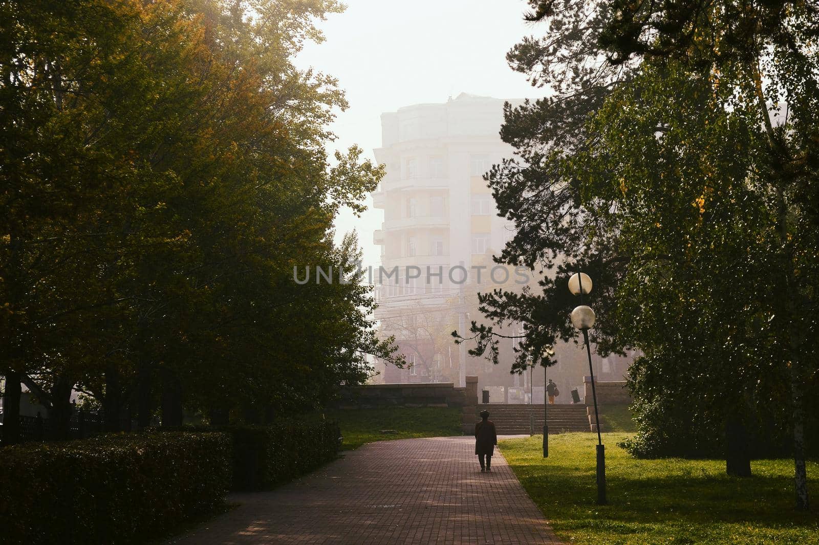 An old woman walks along a stone footpath in the park in the morning among trees, fog and lanterns and a building in the fog in the background by ProjectStockman