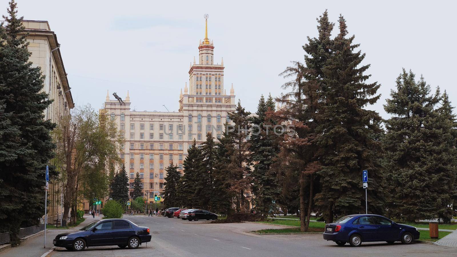 A road and autos and a large number of fir trees with a Soviet building