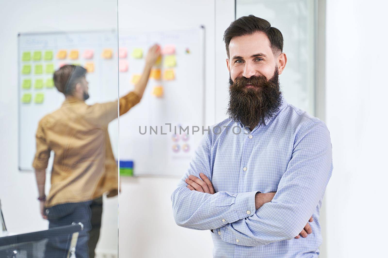 Happy bearded senior developer or manager in modern IT office. Another professional working with scrum desk on background. Successful technology project or startup concept.