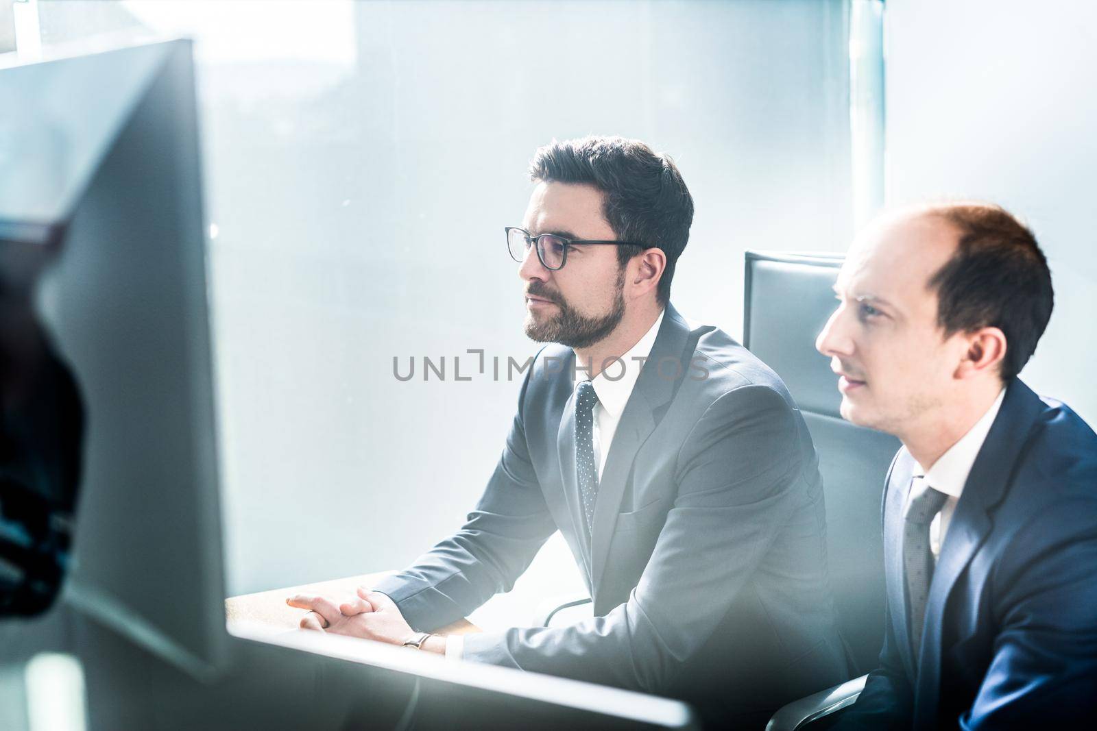 Image of two thoughtful businessmen looking at data on multiple computer screens, solving business issue at business meeting in modern corporate office. Business success concept.