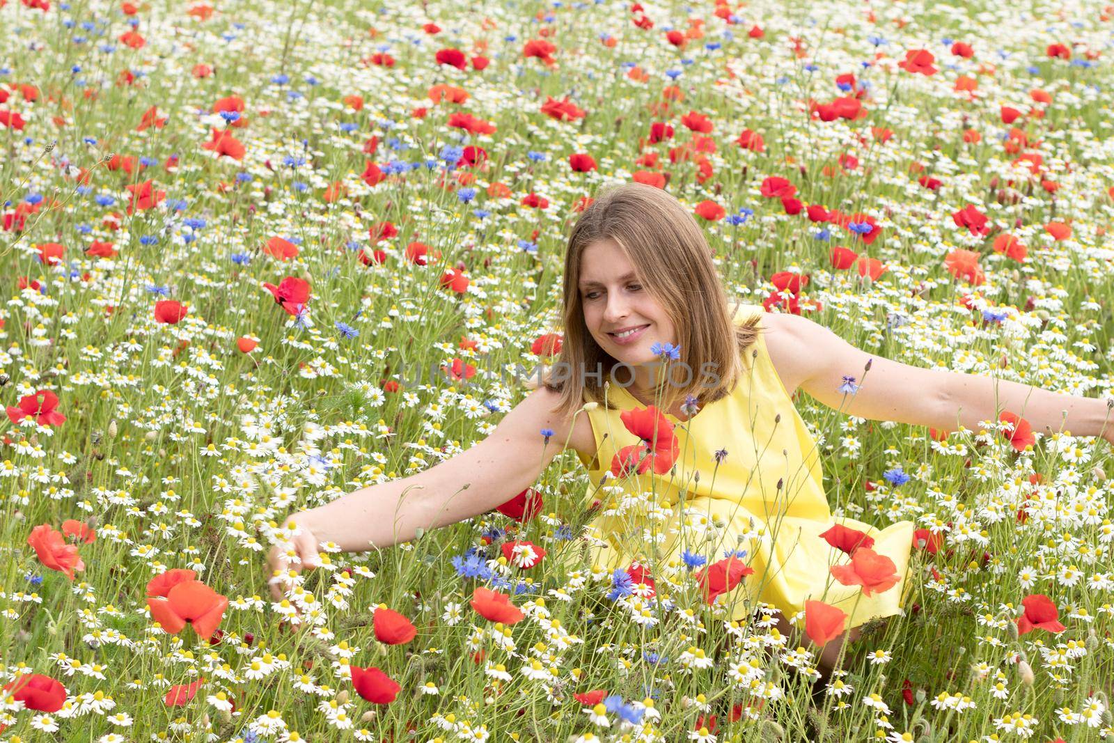 a beautiful young blonde woman in a yellow dress stands among a flowering field of poppies, daisies, cornflowers and laughs. High quality photo