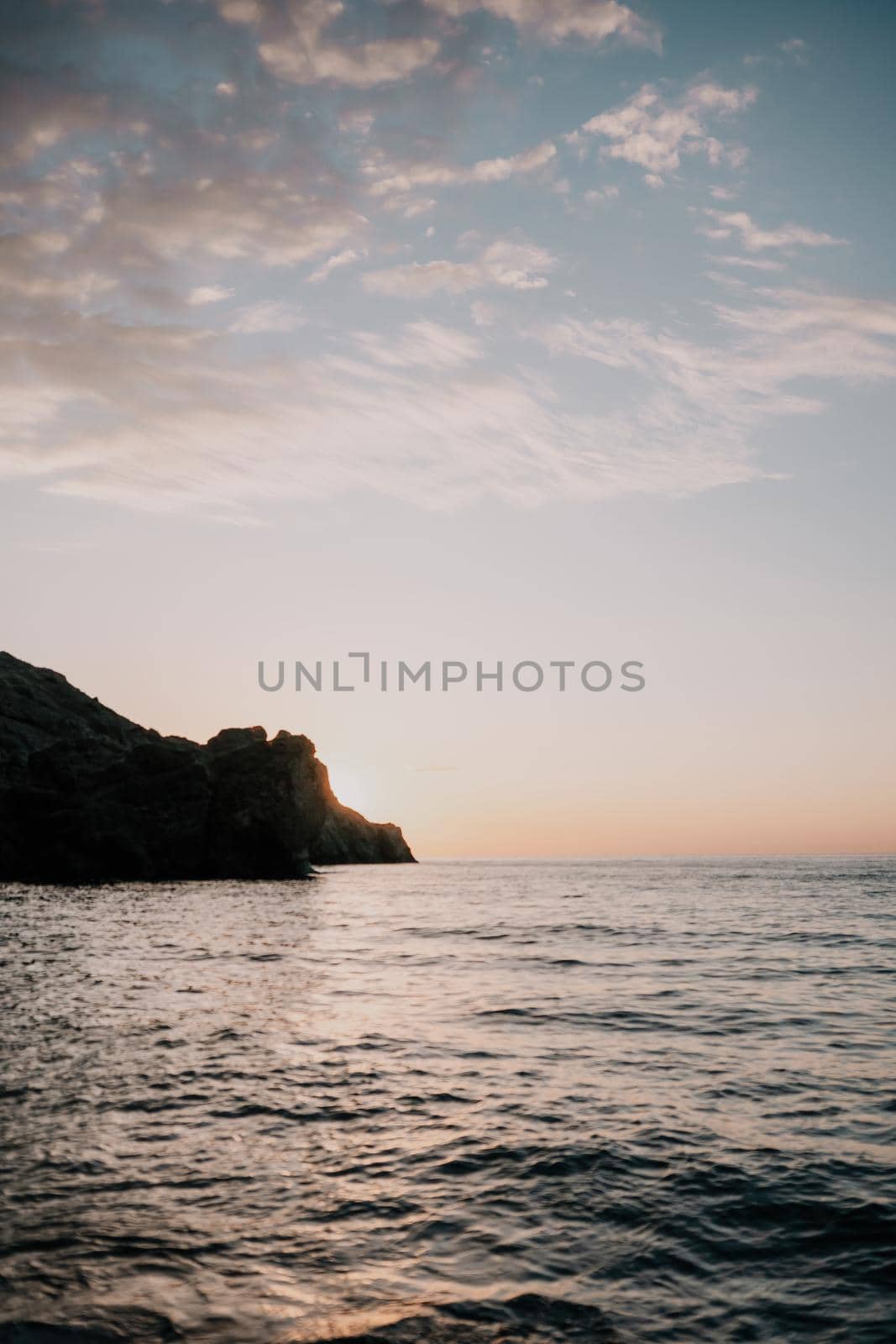 A red burning sunset with the silhouette of a cliff over the sea. Abstract nature summer or spring ocean sea background. Small waves on golden warm water surface with bokeh lights from sun.