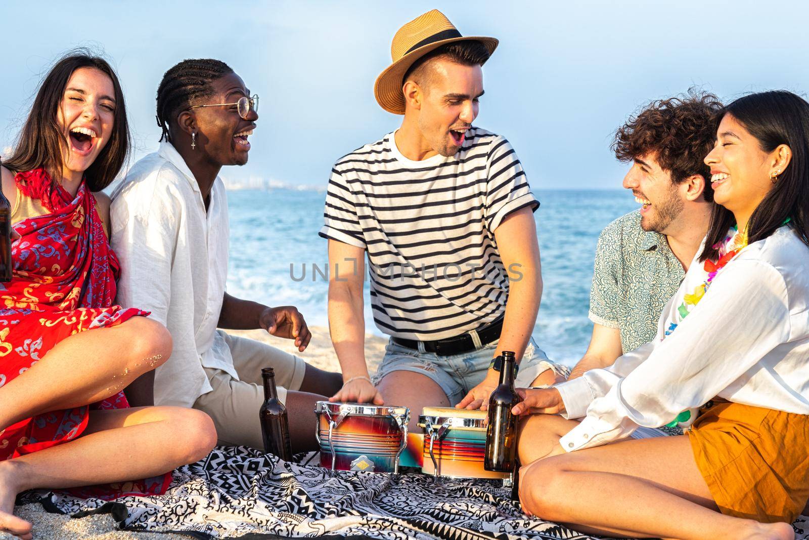 Group of multiracial friends having fun at the beach, laughing out loud, playing bongos and drinking beer together. by Hoverstock