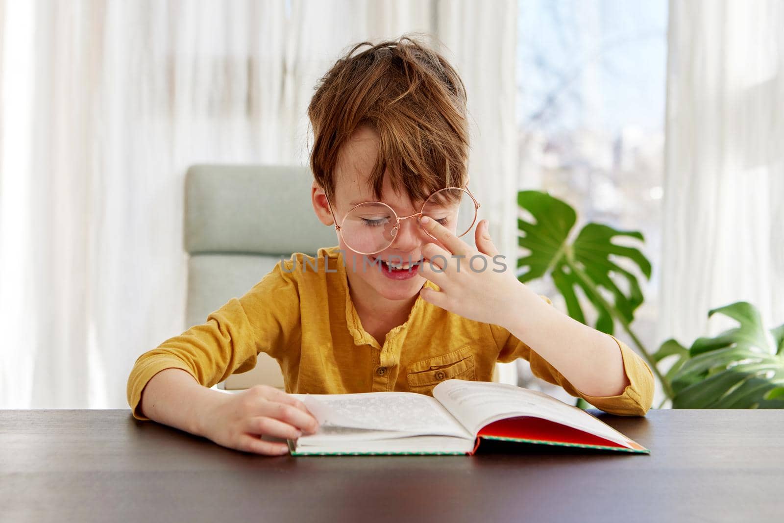 Cute little student in yellow shirt adjusting glasses and smiling while sitting at table and reading book during studies in daytime at home