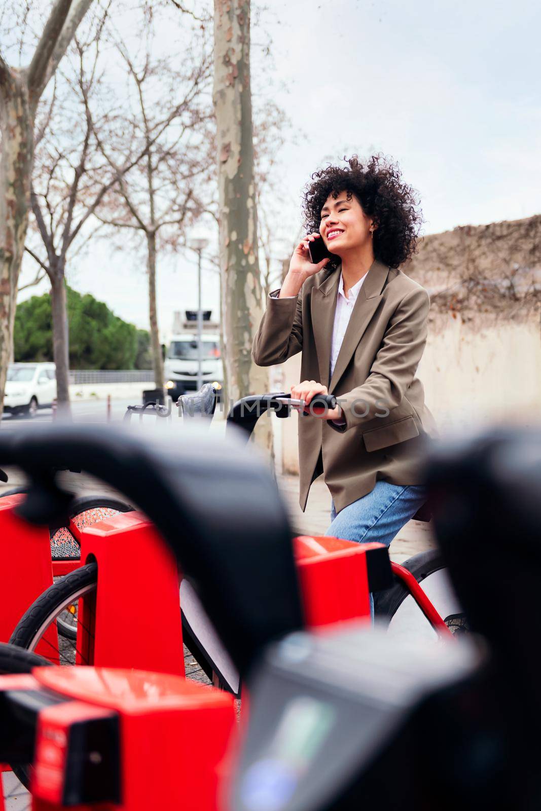 smiling young latin woman talking by phone while sitting on a bike at the rental station, technology and sustainable mobility concept