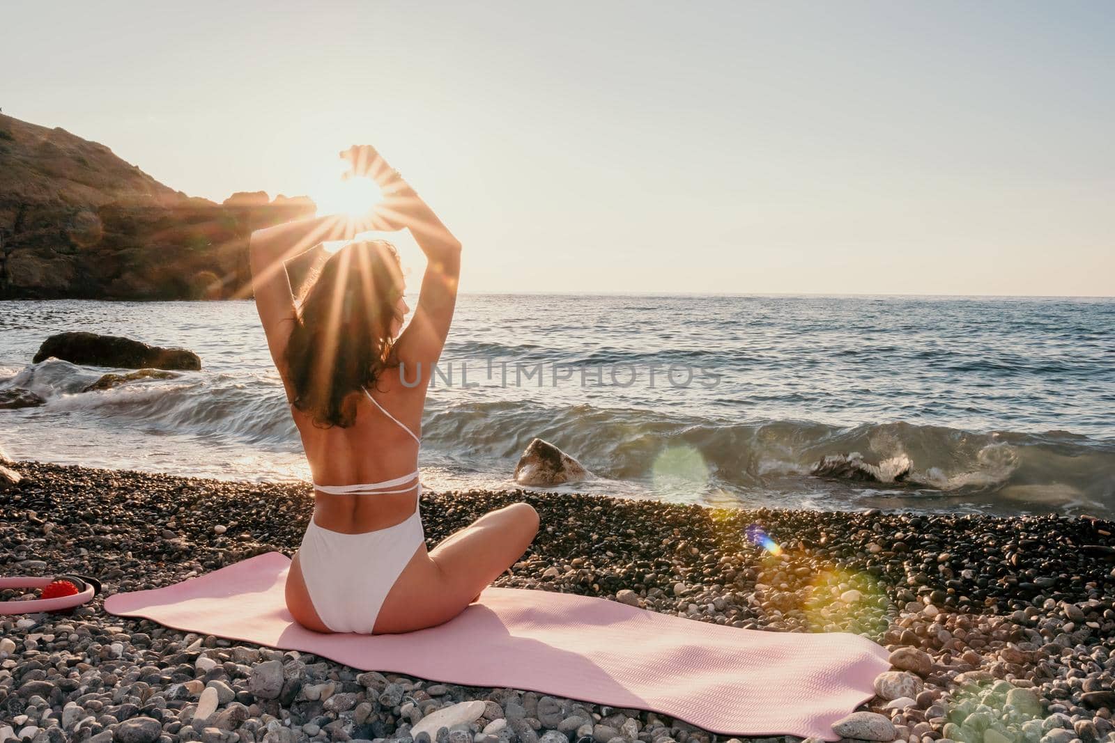 Young woman in swimsuit with long hair practicing stretching outdoors on yoga mat by the sea on a sunny day. Women's yoga fitness pilates routine. Healthy lifestyle, harmony and meditation concept.