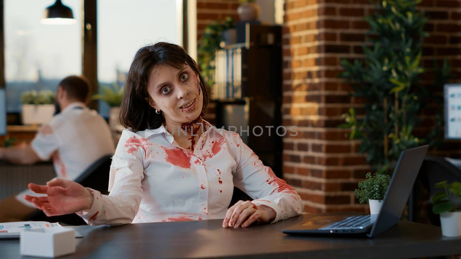 Weird looking spooky zombie woman waving at camera while sitting in office. by DCStudio